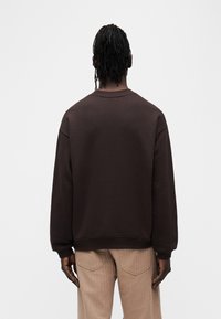 Sweat-shirt en coton marron avec col rond, épaules tombantes et poignets côtelés. Porté avec un pantalon en velours côtelé de couleur claire.