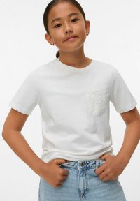 Camiseta de algodón blanca con mangas cortas, escote redondo y detalle de bolsillo bordado intrincado. Combinada con pantalones cortos de mezclilla azul claro.