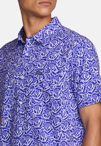 Short-sleeve button-up shirt made of blue fabric with a white rose pattern; features a collar and a small RVCA logo patch.