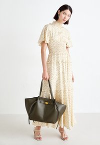 Kermanvärinen mekko, jossa on tummat pallokuvioinnit, röyhelöhihat ja smokkivyötärö. Oliivinvihreä nahkainen olkalaukku, jossa on kullanvärinen lukko, seisoo valkoisella taustalla.