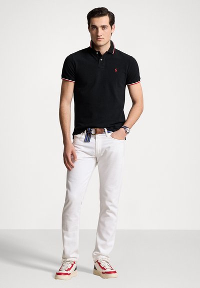 Black polo shirt with red and white accents, paired with white jeans and red, white, and blue sneakers. Model displays a casual outfit.