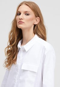 White cotton button-up shirt with a pointed collar, single chest pocket, and subtle sheen, featuring a straight cut and long sleeves.