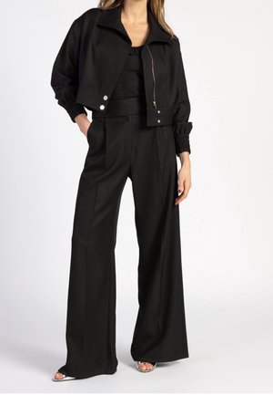 Frau trägt eine schwarze, kurz geschnittene Jacke mit Reißverschluss und Knöpfen, kombiniert mit hoch taillierten, weit geschnittenen schwarzen Hosen und silbernen offenen High Heels.