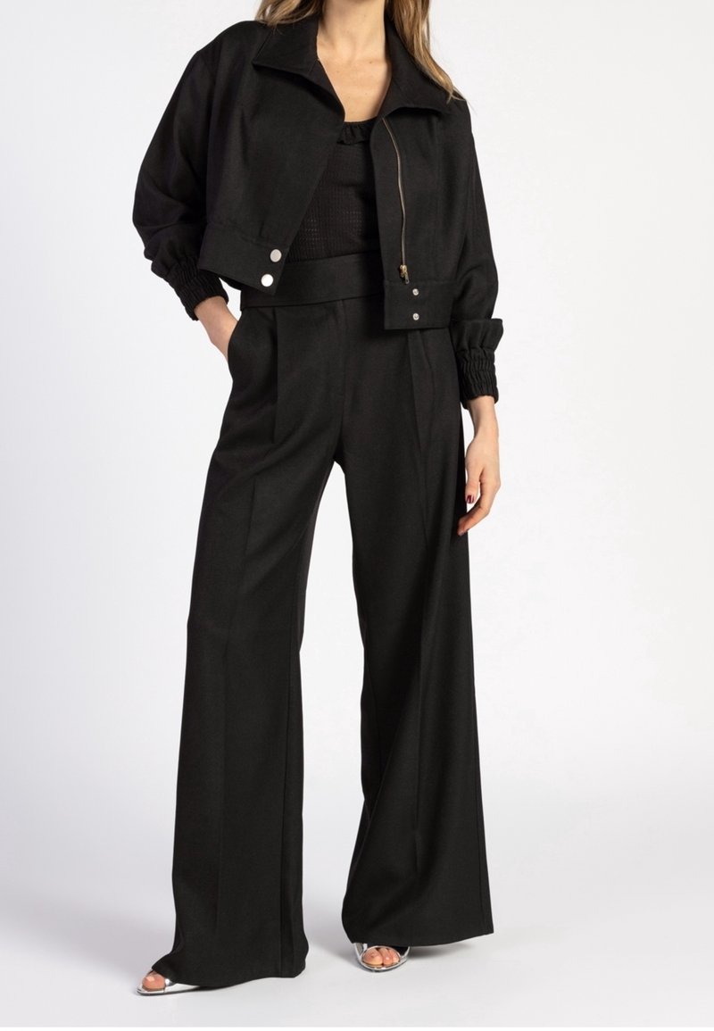 Femme portant une veste courte noire avec fermeture éclair et boutons, associée à un pantalon noir taille haute à jambes larges et des talons ouverts argentés.