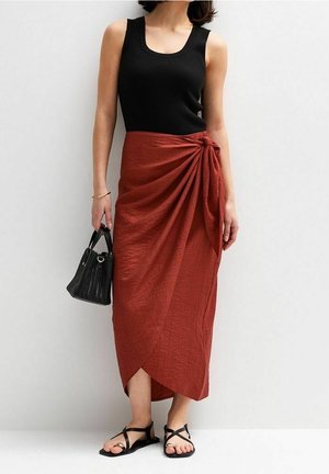 Femme portant un haut noir sans manches, une jupe portefeuille couleur rouille avec un détail noué, des sandales noires et tenant un sac à main noir.