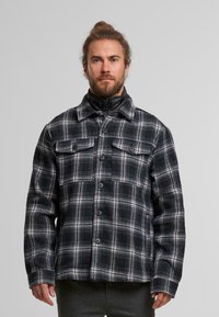 Schwarze und weiße kariierte Flanelljacke mit Knopfverschluss, zwei Brusttaschen und hohem Kragen. Die Textur wirkt weich und warm.