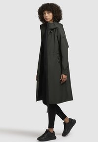 Cappotto impermeabile lungo, di un verde scuro, con cappuccio, chiusura con zip, vita regolabile con cordino e tasche laterali. Indossato sopra leggings neri e sneakers.