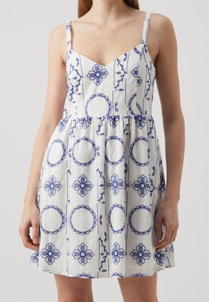 Witte mouwloze jurk met dunne bandjes, met blauwe bloem- en cirkelborduursels, gedragen door een staande persoon.