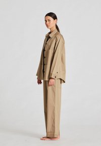 Beige oversized paita, jossa on mustat napit ja taskut, yhdistettyinä samanvärisiin leveälahkeisiin housuihin. Tasainen kangasrakennus, minimalistinen muotoilu.