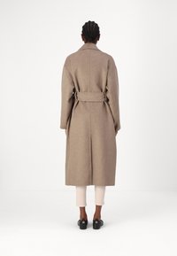 Long manteau en laine beige avec une taille ceinturée, de larges revers et une coupe droite, associé à un pantalon ajusté de couleur claire et des chaussures noires.