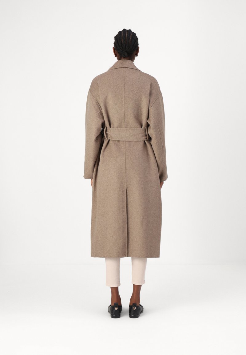 Langer, beiger Wollmantel mit einem Gürtel in der Taille, breiten Revers und gerader Schnitt, kombiniert mit hellen, taillierten Hosen und schwarzen Schuhen.