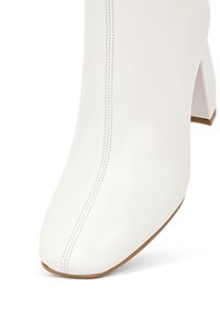Bottine en cuir blanc, à bout pointu, avec des coutures relevées et un talon bloc épais. Texture lisse et design minimaliste.
