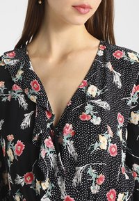 Mustikka kukkakuosiset pusero, jossa on V-pääntie, pilkkukuvio, röyhelökaulus ja elävät kukkakuosit punaisina, vaaleanpunaisina ja keltaisina. Kevyt kangas.