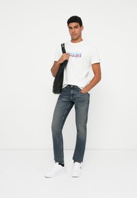 Άνδρας σε casual ντύσιμο, φορώντας λευκό γραφικό T-shirt, μπλε τζιν, λευκά αθλητικά παπούτσια, κρατώντας μαύρη τσάντα ώμου σε απλή φόντο.