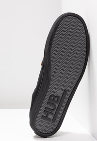 Semelle noire de basket avec motif ondulé, nom de la marque "HUB" embossé près du talon, chaussure inclinée contre un mur blanc.