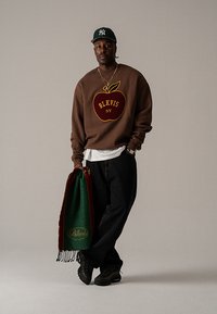 Hnedá mikina s motívom jablka a textom "BLKVIS NY". Čierne džínsové nohavice a zeleno-červeno strapcová šatka. Čierne tenisky. Zelená šiltovka.