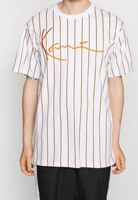 Camiseta blanca con rayas verticales naranjas y una firma bordada en naranja en el frente. Mangas cortas, corte relajado, material de algodón.