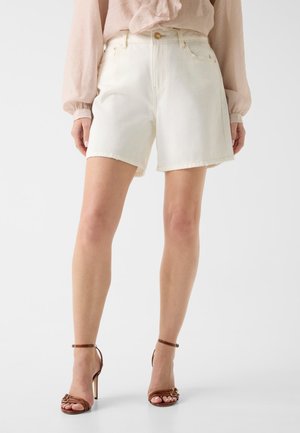Frau trägt cremefarbene High-Waist-Denim-Shorts kombiniert mit braunen High-Heel-Sandalen und einer hellrosa Bluse.