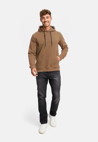 Brun hættebluse med en tekstureret finish, der har en kangaroo lomme og sorte snore. Bæret med mørke jeans og hvide sneakers.