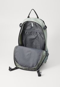 Rucksack aus gedämpftem grünem und grauem Stoff, mit einem großen Hauptfach mit Klappe, Netzdetails und verstellbaren Trägern.