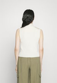 Top senza maniche in beige chiaro, design aderente con scollatura alta. La parte posteriore presenta un orlo discreto e una finitura testurizzata.