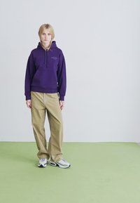 Nuori henkilö, jolla on vaaleat hiukset, pukeutuneena violettiin huppariin, vaaleisiin housuihin ja valkoisiin lenkkareihin, seisoo vaaleanvihreällä lattialla valkoista seinää vasten.