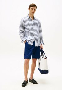 Gestreept blauw-wit shirt met lange mouwen, gecombineerd met blauwe shorts. Zwarte instappers en een witte tote bag met marineblauwe accenten.