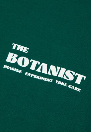 Un tessuto di cotone verde presenta un bold testo bianco che recita "THE BOTANIST" con le parole "IMAGINE EXPERIMENT TAKE CARE" in lettere più piccole sottostanti.
