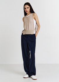 Gilet lavorato a maglia beige con lacci laterali, indossato sopra una maglietta bianca. Abbinato a pantaloni a gamba larga a righe navy e sneakers bianche.