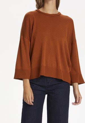Femme portant un pull en maille rouille ample à manches trois-quarts et un jean bleu foncé, se tenant contre un fond blanc uni.