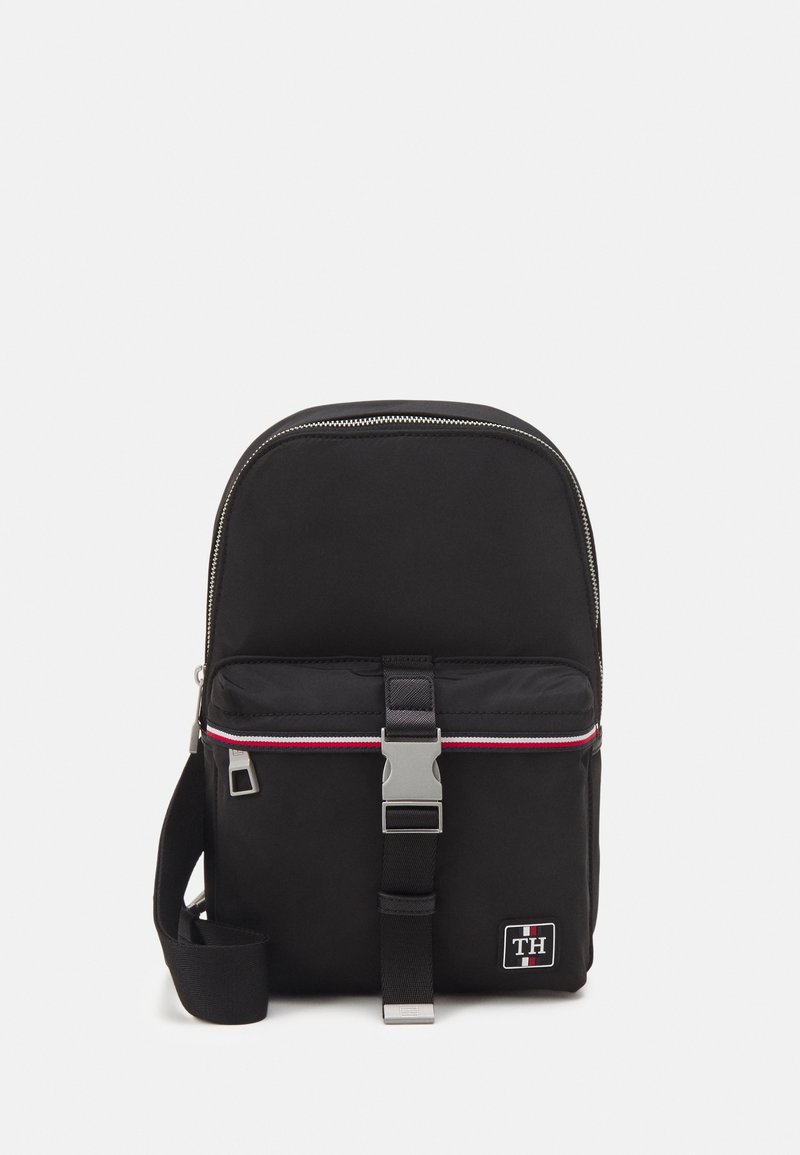 Zaino in nylon nero con una parte superiore arrotondata, zip argentata, striscia orizzontale rossa e bianca, tracolla regolabile e tasca frontale con una chiusura in metallo.