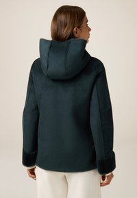 Chaqueta con capucha de color verde oscuro, hecha de una tela suave y lisa, con puños y capucha forrados de pelo. Tiene un corte relajado y un diseño minimalista.