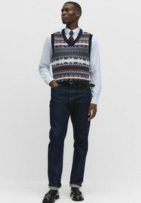 Marinblå och vit mönstrad stickad väst över en ljusblå skjorta, i kombination med mörka jeans och svarta skor. Har en V-ringad design.