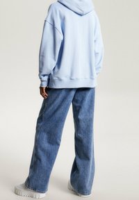 Persona vistiendo una sudadera azul clara, jeans holgados azules y zapatillas deportivas blancas de suela gruesa, de pie contra un fondo claro y liso.
