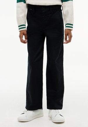 Person iført sorte bukser med lige ben, hvide sneakers og en hvid sweater med grønne striber på ærmerne, stående mod en hvid baggrund.