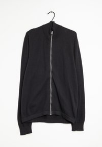 Svart zip-up jacka med ett strukturerat mönster, hög krage och långa ärmar. Tillverkad av ett mjukt, stickat material. Hänger på en krok.