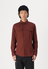 Camisa castanha de mangas longas, feita de tecido texturizado, com colarinho abotoado, dois bolsos no peito e botões pretos.