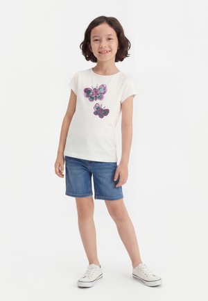 Nuori tyttö hymyilee, hänellä on valkoinen t-paita, jossa on kaksi paljetti-perhosta, farkkushortsit ja valkoiset tennarit, seisoo valkoista taustaa vasten.