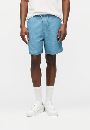 Hombre vistiendo pantalones cortos de mezclilla azul claro con cordón, calcetas blancas hasta la pantorrilla, zapatillas deportivas blancas y una camiseta blanca, de pie contra un fondo blanco.