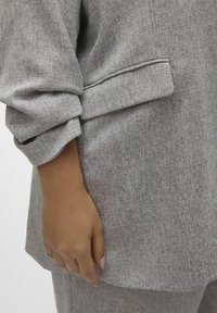 Grijze textuurjacket met opgerolde mouwen, met een zijzak, weergegeven in close-up naast een hand in een natuurlijke positie.