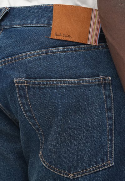 Sinised teksapüksid, millel on helepruun nahast etikett mitmevärviliste triipudega, mille ülemises vasakus nurgas on reljeefna "Paul Smith".