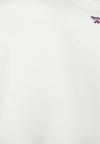 Tessuto bianco Reebok con una piccola toppa logo intessuta in bianco, rosso e nero cucita vicino all'angolo in alto a destra.