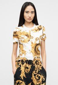 Νεαρή γυναίκα με μακριά σκούρα μαλλιά φοράει ταιριαστό λευκό και μαύρο σύνολο με περίπλοκα χρυσά σχέδια μπαρόκ με στροβιλισμούς.