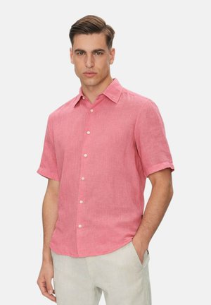 Mann trägt ein korallenrosa kurzärmeliges Hemd mit Knöpfen und helle beige Hose, steht mit einer Hand in der Tasche vor weißem Hintergrund.