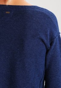 Pull en maille bleu marine avec un large décolleté, présentant une surface texturée et une petite étiquette métallique avec logo à l'arrière.