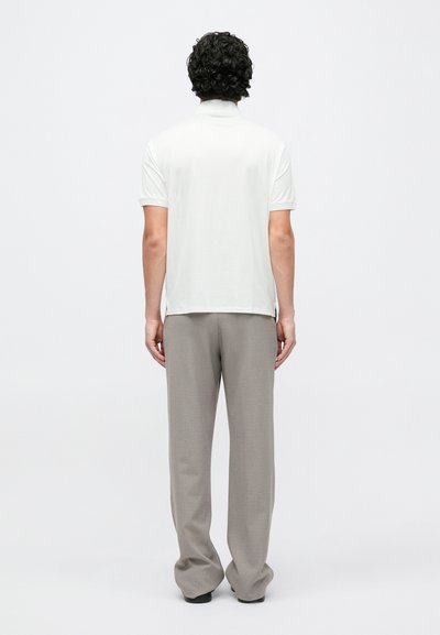 Pánské bílé polo tričko s krátkými rukávy, volnější střih, boční rozparky, v kombinaci se světle šedými kalhotami. Hladká textura, jednoduchý design.