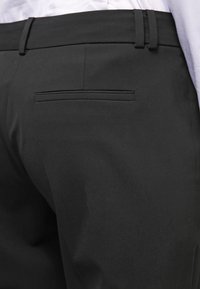 Pantalon noir en tissu lisse, avec une poche arrière plate et une taille cintrée dotée de passants de ceinture. Design épuré et minimaliste.