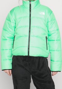 Person iført en lys grøn polstret jakke med sort lynlås og The North Face-logo, kombineret med sorte teksturerede bukser.