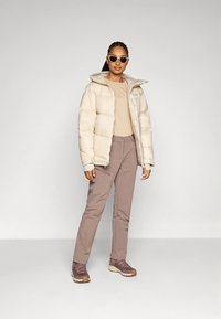 Donna che indossa una giacca a vento beige, una maglietta a maniche lunghe color tabacco, pantaloni malva, sneakers marroni e occhiali da sole bianchi, in piedi su uno sfondo neutro.