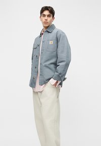 Νέος άνδρας που φοράει ανοιχτό μπλε εργασιακό μπουφάν πάνω από ροζ πουκάμισο και κρεμ παντελόνι με φαρδιά μπατζάκια, στέκεται μπροστά σε λευκό φόντο.